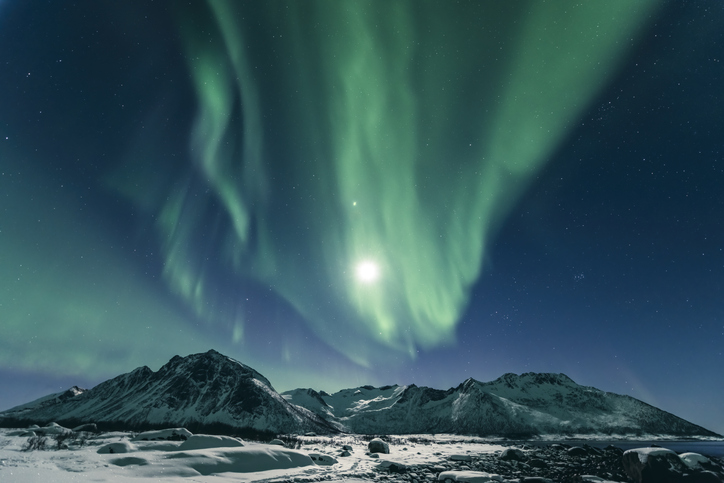 Northern Lights, polar light or Aurora Borealis in the night sky over Senja island in Northern Norway. Snow covered mountains in the background are illuminated by moonlight in a snow covered landscape.