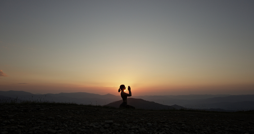 A tranquil moment captured as a young woman practices yoga in prayer position, silhouetted against the rising sun on a mountain summit.